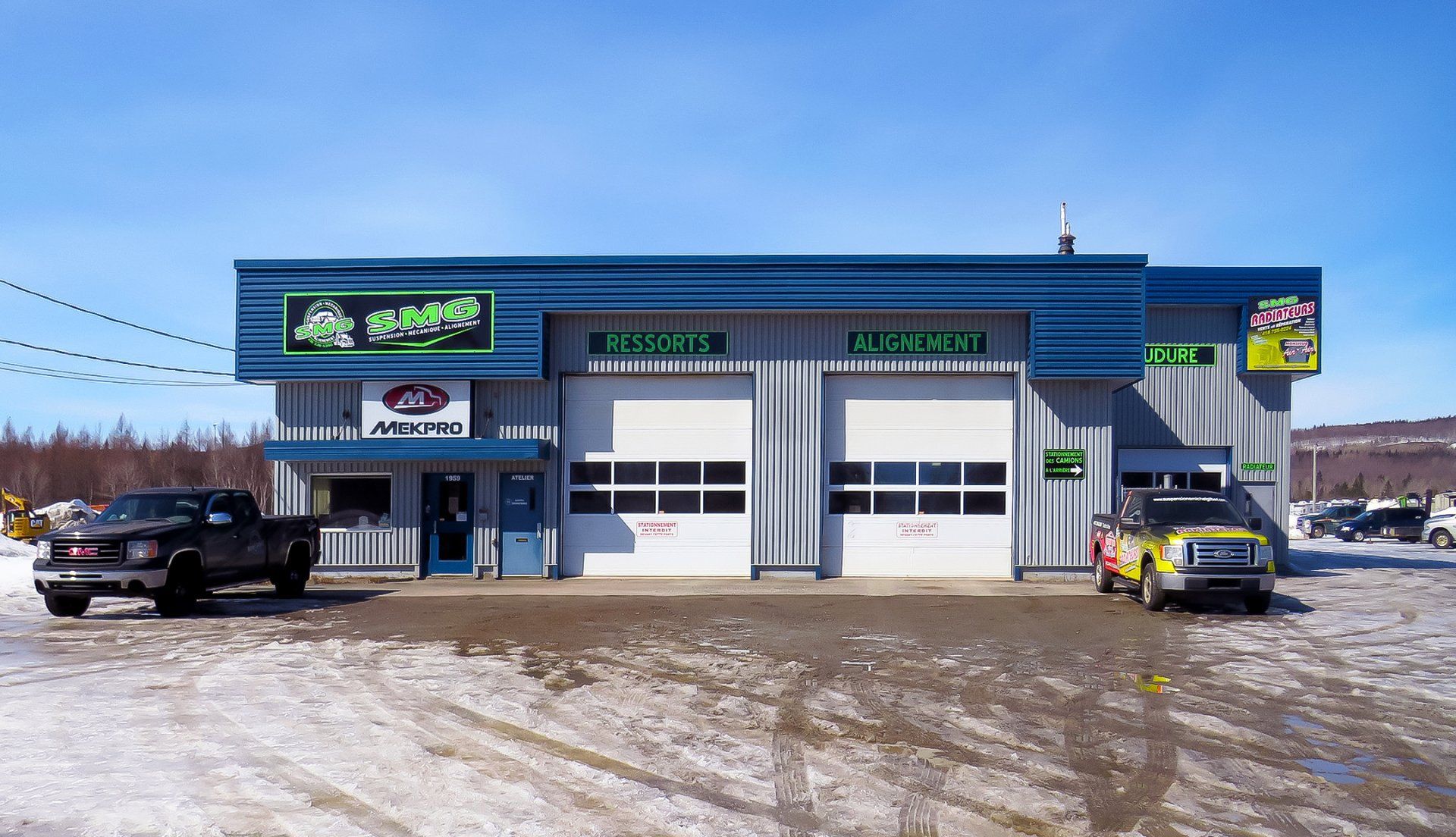Atelier de réparation automobile avec deux portes de garage, un camion noir et un camion jaune garés à l'extérieur, neige au sol.