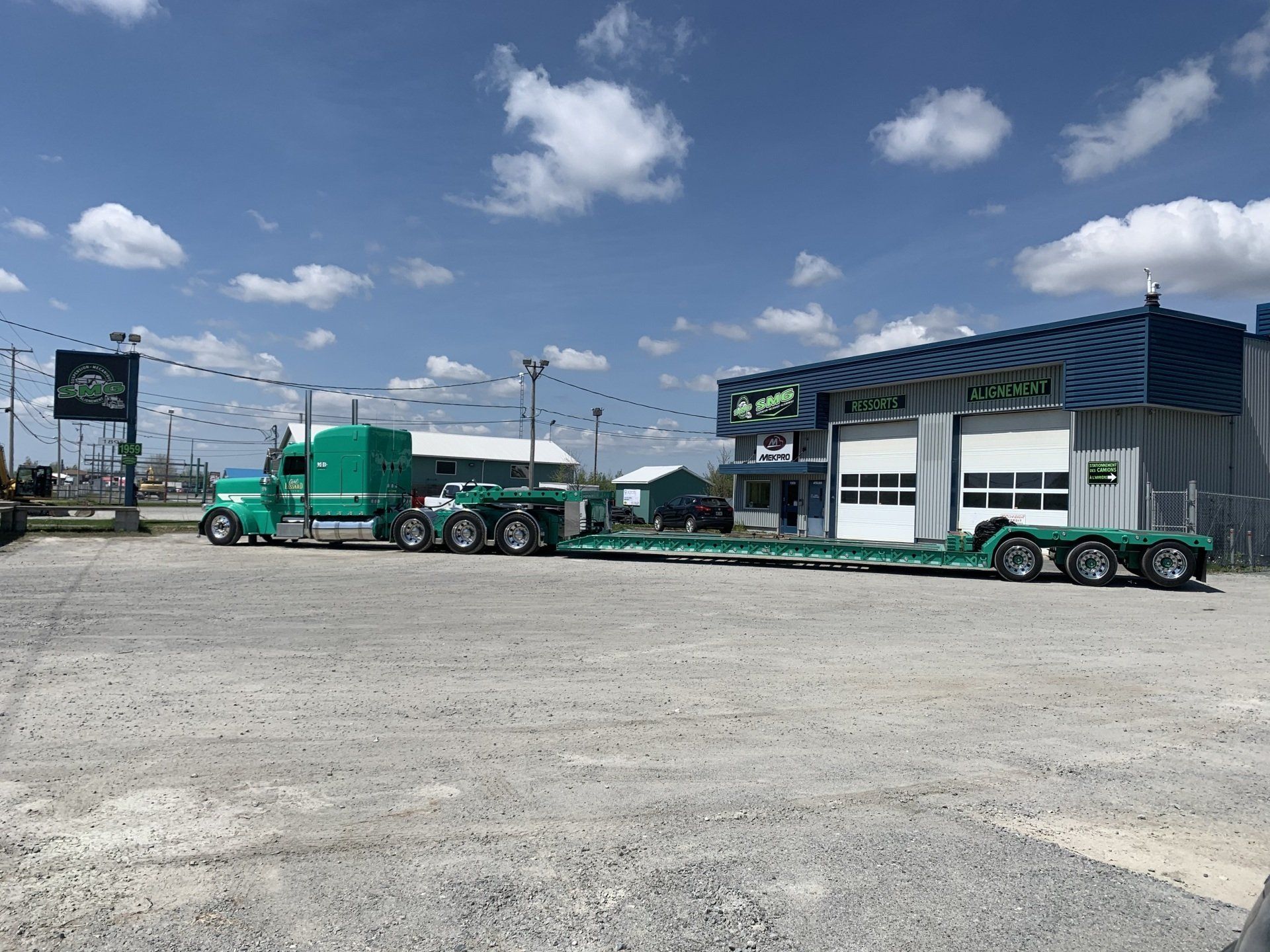 Semi-camion vert avec une longue remorque stationné devant une entreprise par une journée ensoleillée.