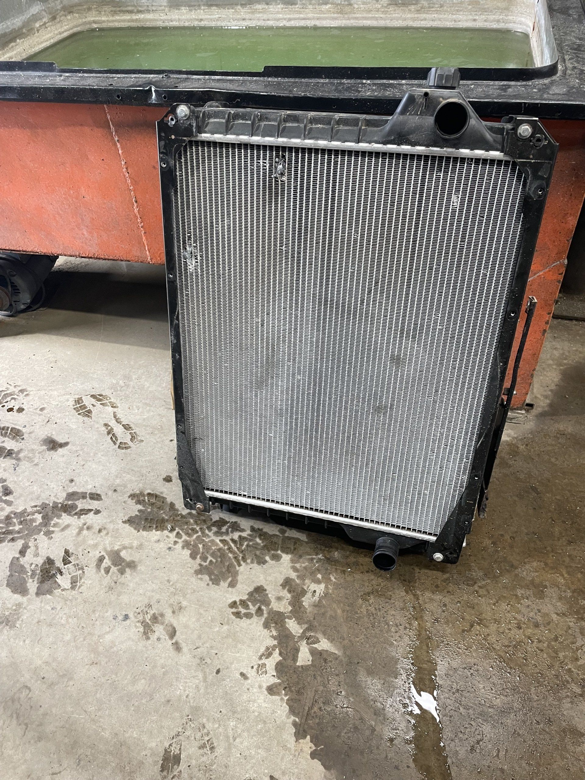 Radiateur de voiture avec ailettes métalliques, cadre noir, sur une surface en béton humide près d'un évier industriel marron.