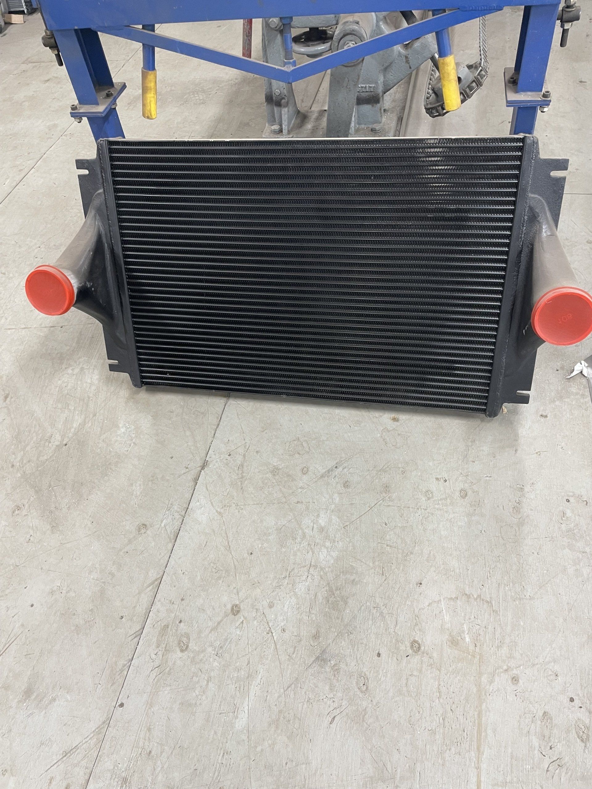 Intercooler noir avec bouchons rouges, devant une structure de support bleue sur un sol en béton.