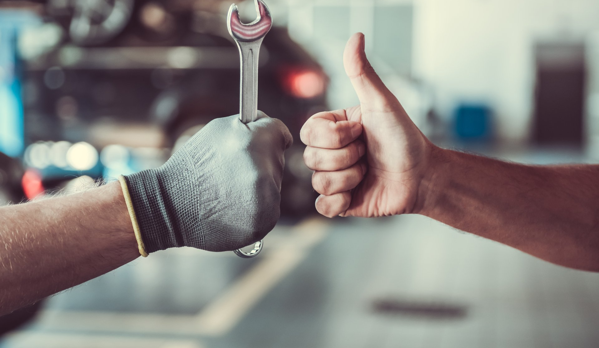 Deux mains, l'une tenant une clé, l'autre levant le pouce dans un atelier de réparation automobile.