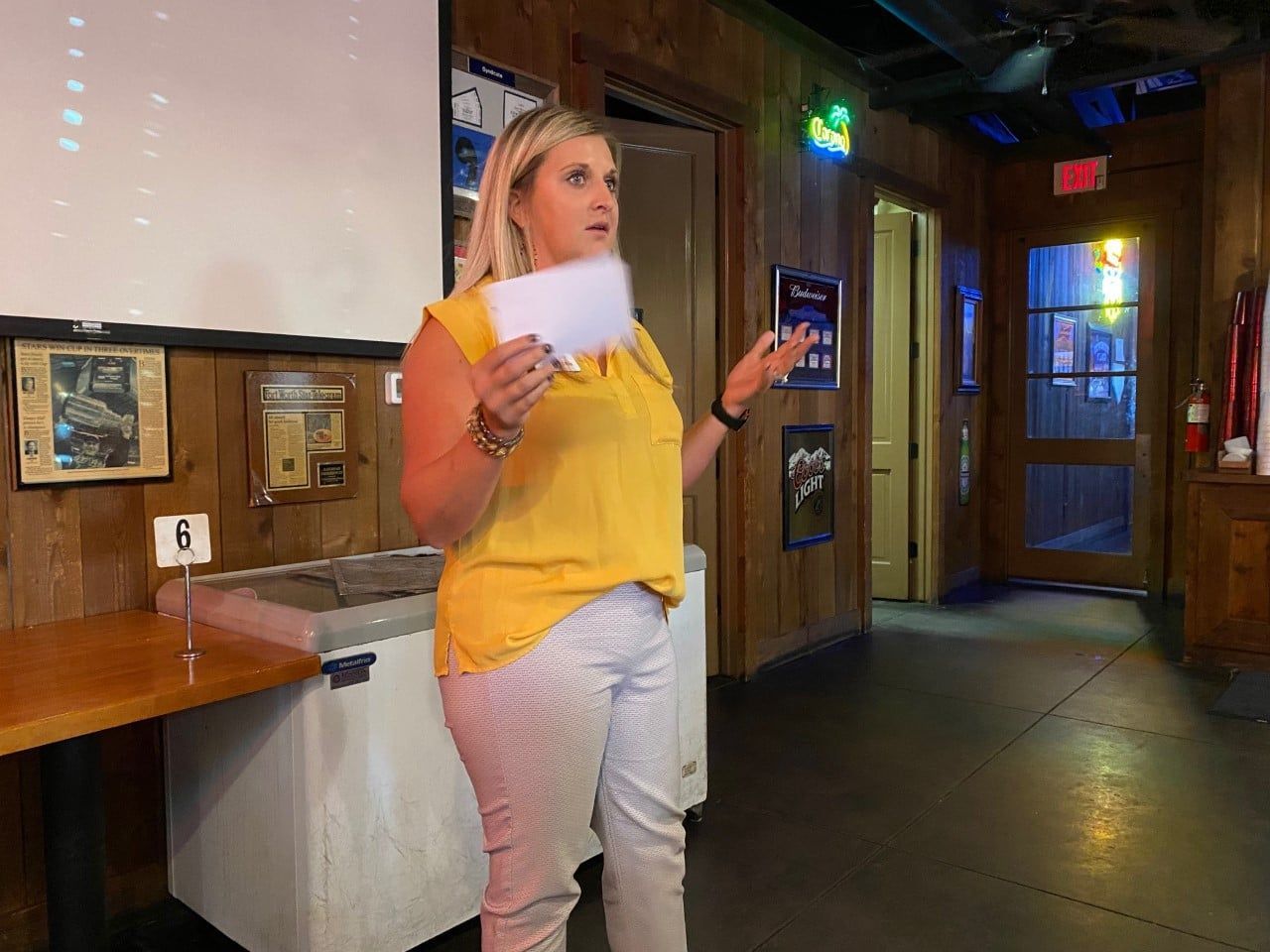 A woman is standing in front of a projector screen in a room, holding a piece of paper.