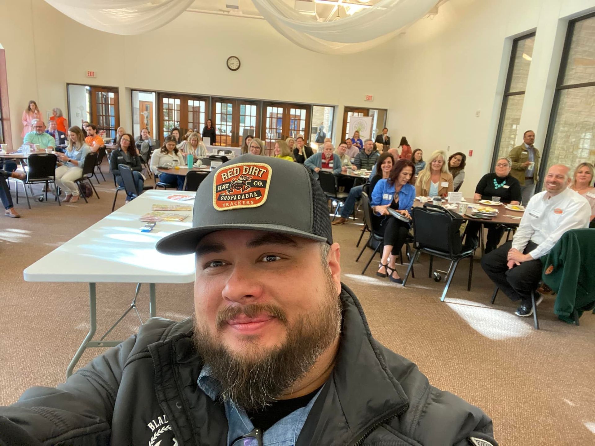 A man with a beard is taking a selfie in front of a group of people sitting at tables.