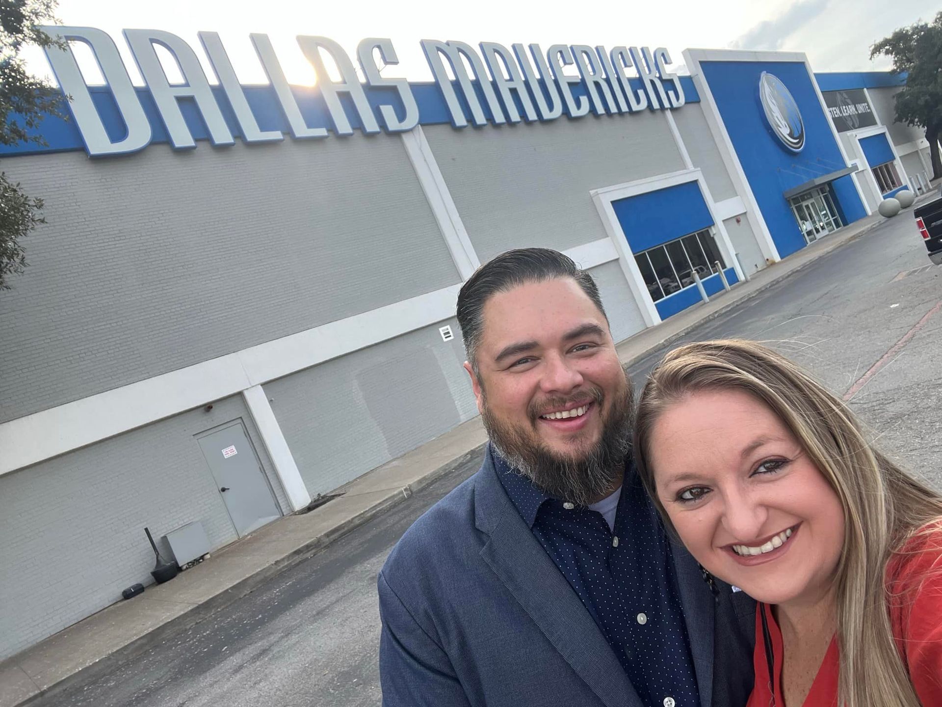 A man and a woman are posing for a picture in front of a Dallas Mavericks building.