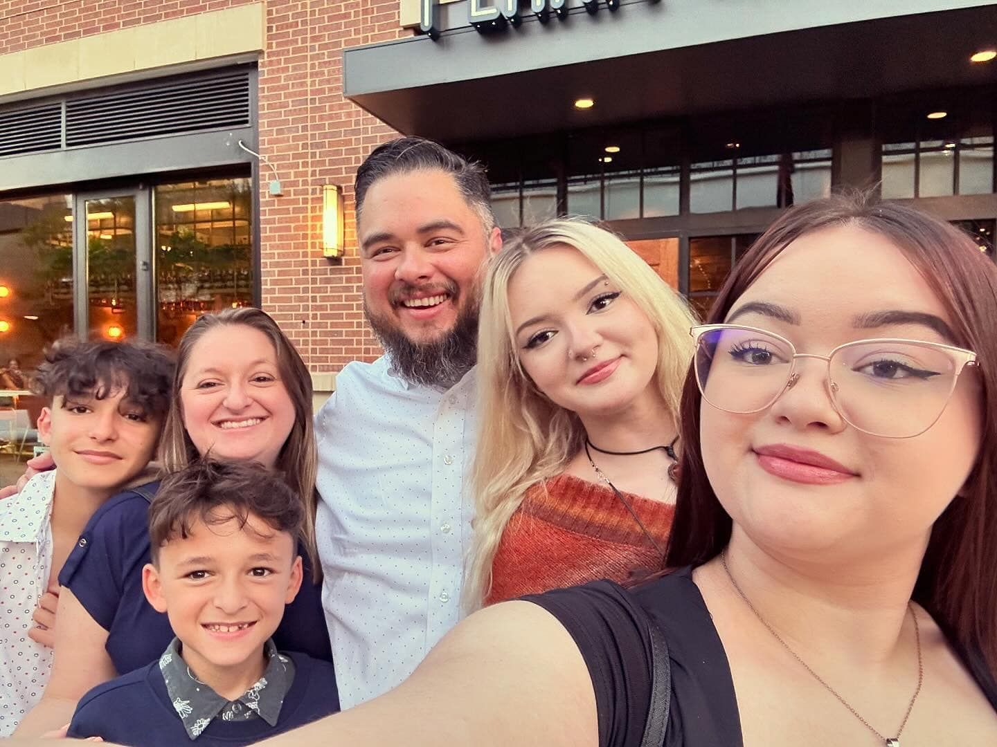 A family of six smiles outdoors in front of a brick building, taken from a selfie angle.