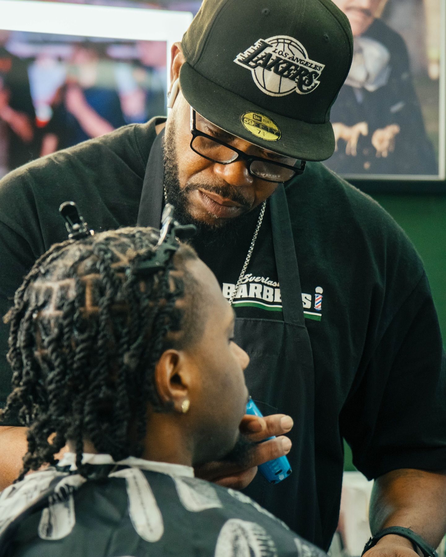 Barber wearing a Lakers cap cutting a client's hair in a shop, holding clippers.