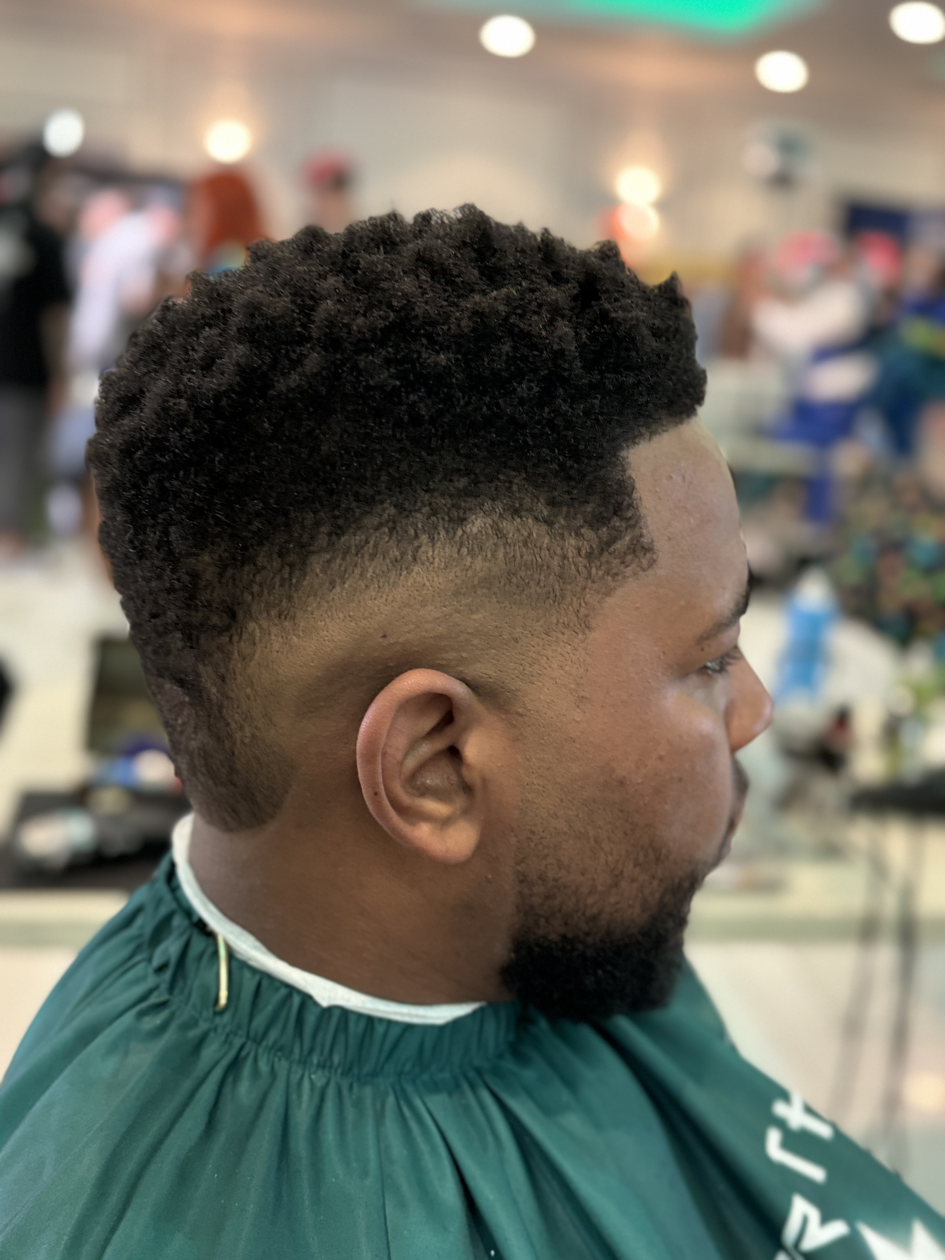 Man with a faded haircut, dark textured hair on top, wearing a green barber cape; indoors.