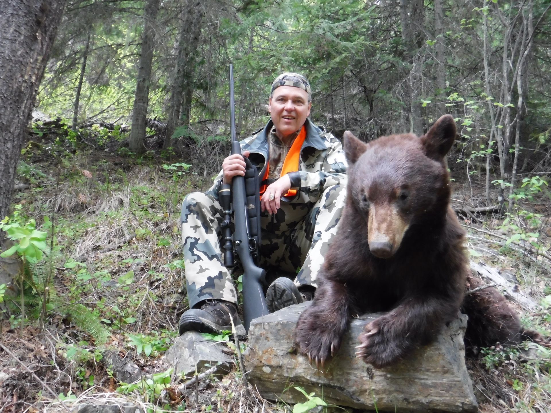 A man is sitting next to a black bear in the woods.