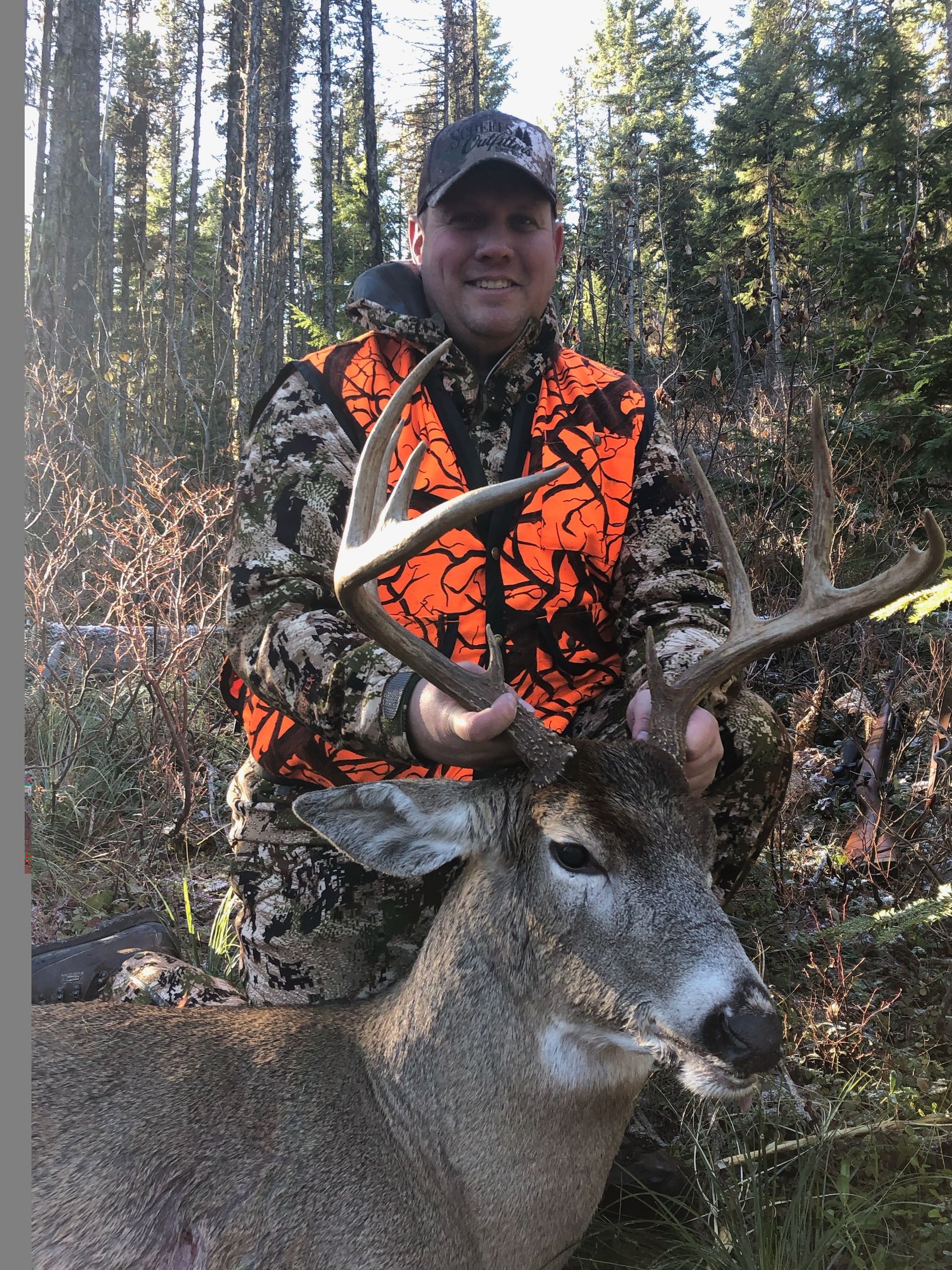 A man in an orange vest is holding a deer in the woods.