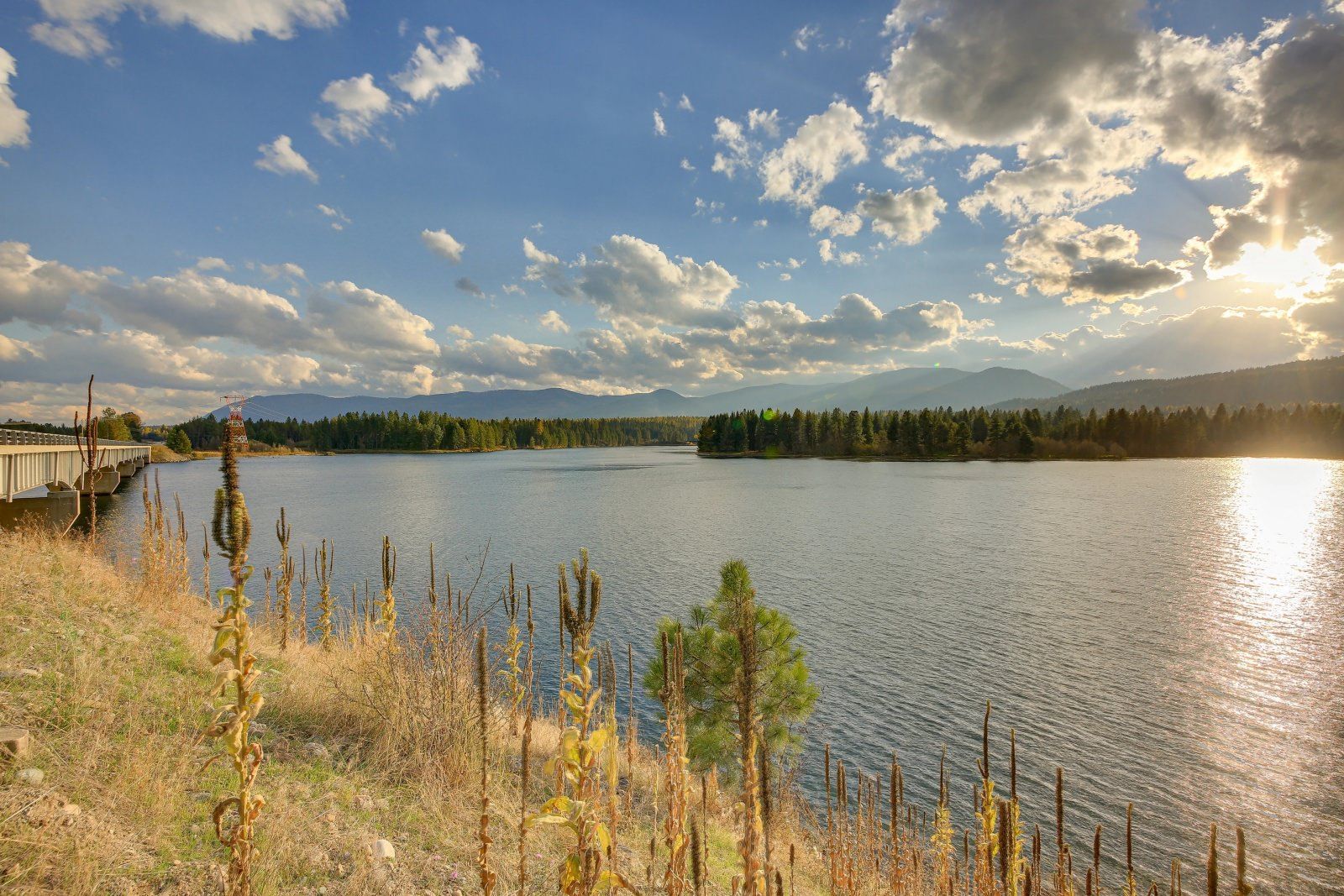A large body of water with mountains in the background and the sun shining through the clouds.
