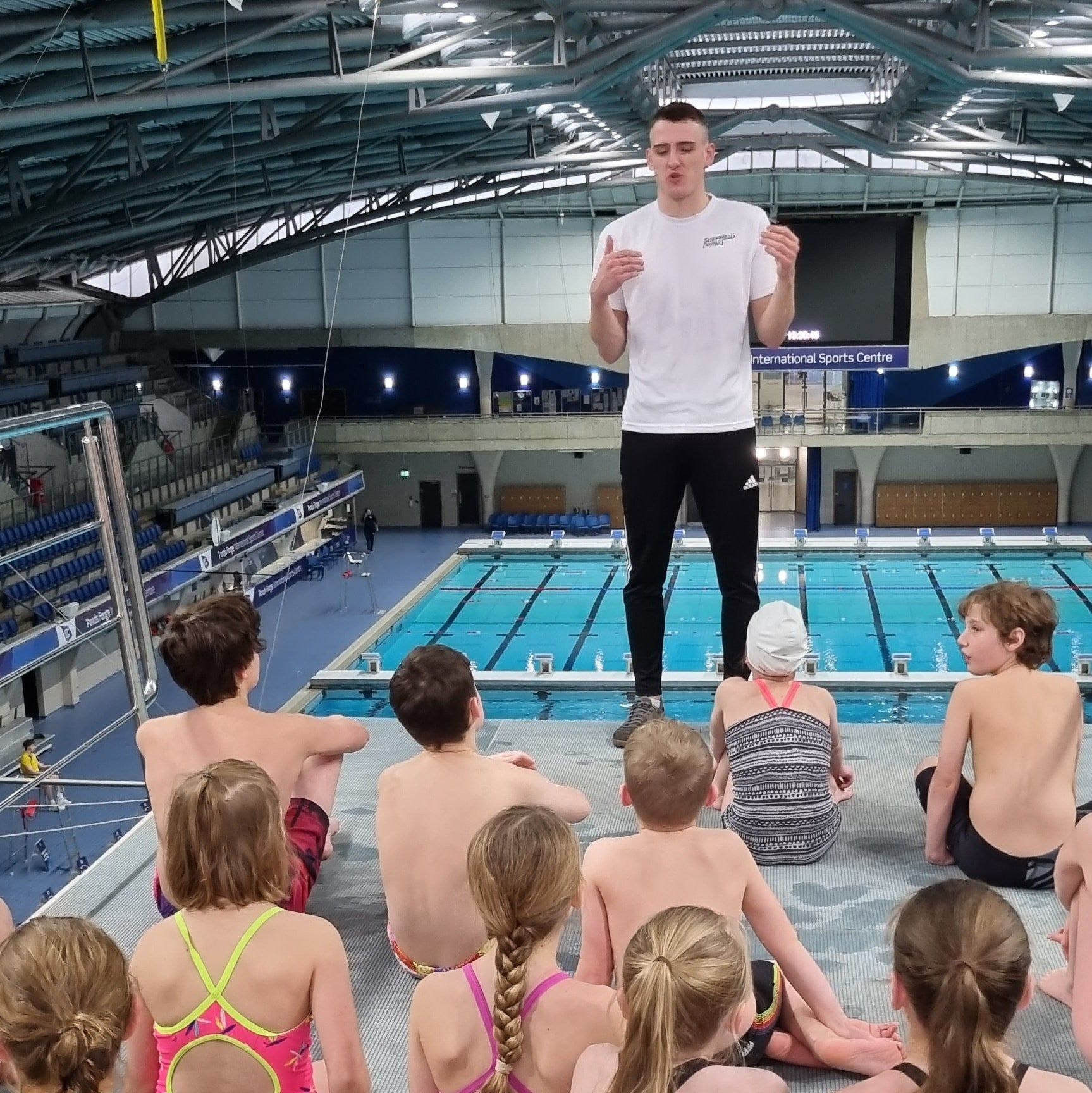 A man is giving a presentation to a group of children in front of a swimming pool