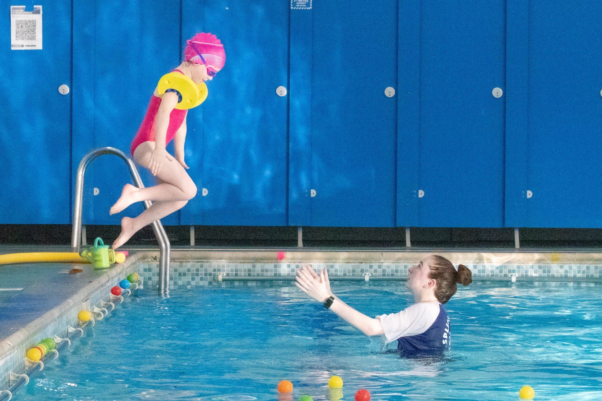 A little girl is jumping into a swimming pool while a woman watches.