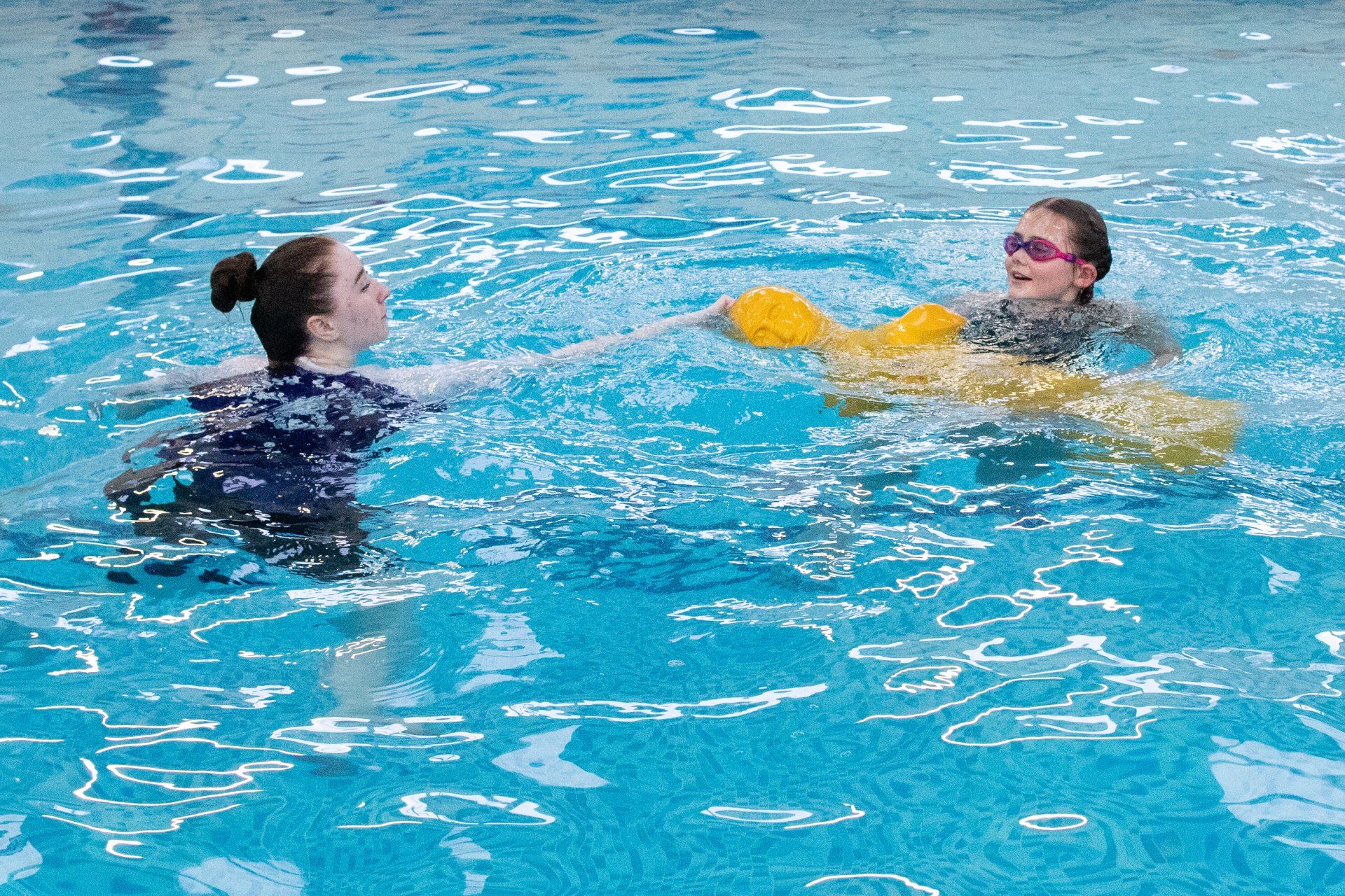 Two women are swimming in a pool with a yellow life preserver.