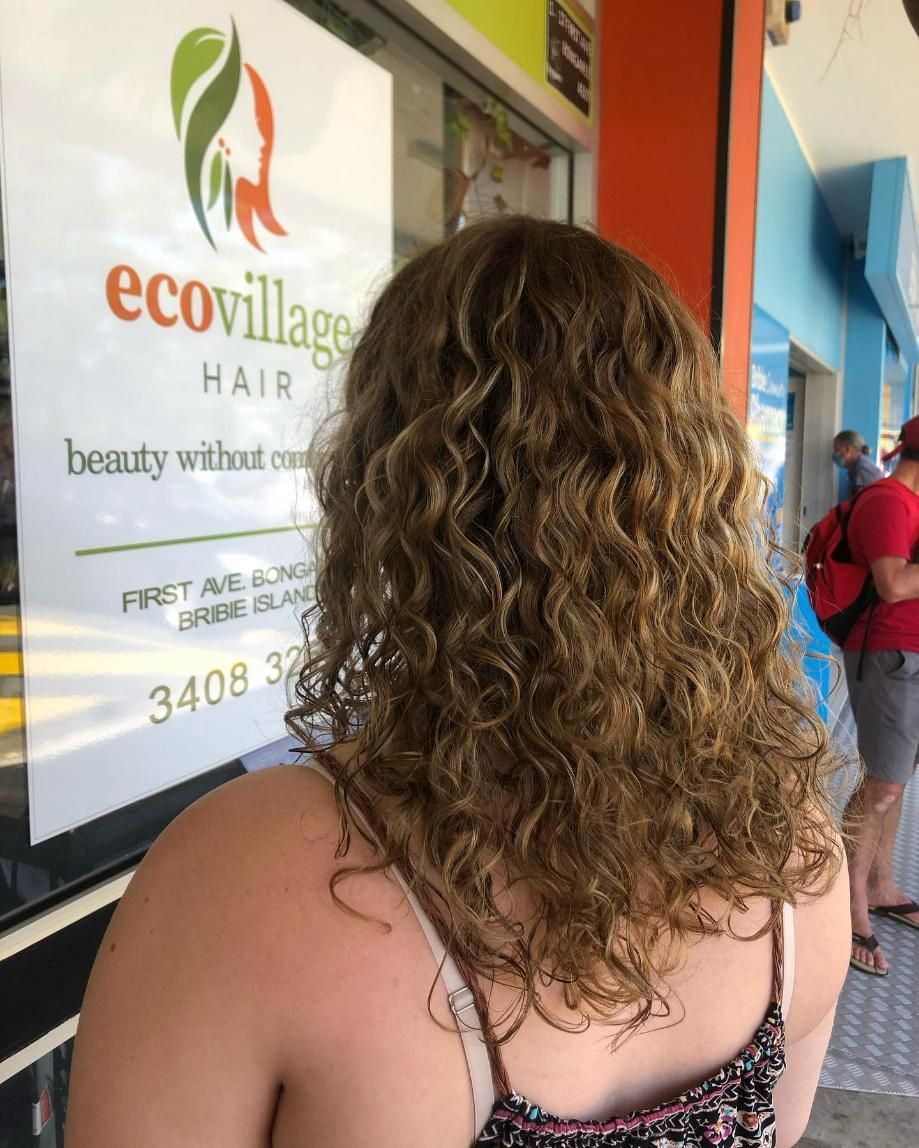 A Woman with Curly Hair Is Standing in Front of A Sign for Eco Village Hair — Eco Village Hair in Bongaree, QLD