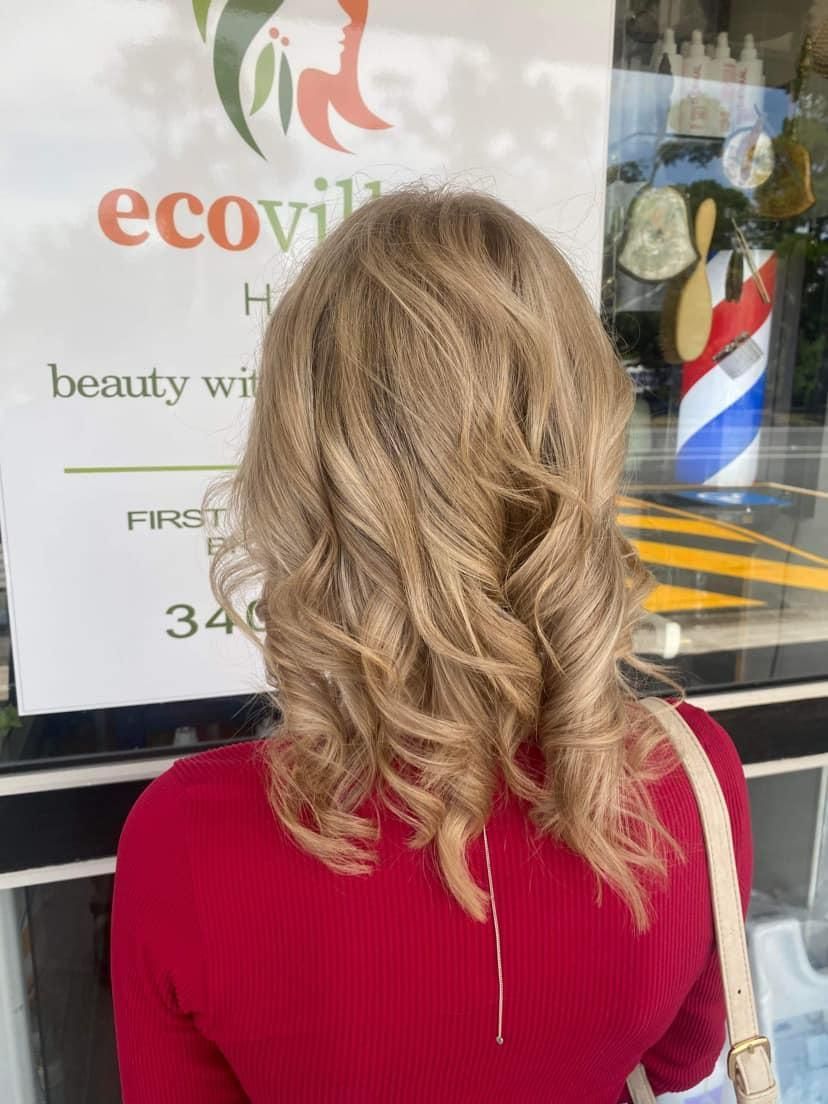 A Woman with Blonde Hair Is Standing in Front of A Sign that Says Eco Village Hair — Eco Village Hair in Bongaree, QLD