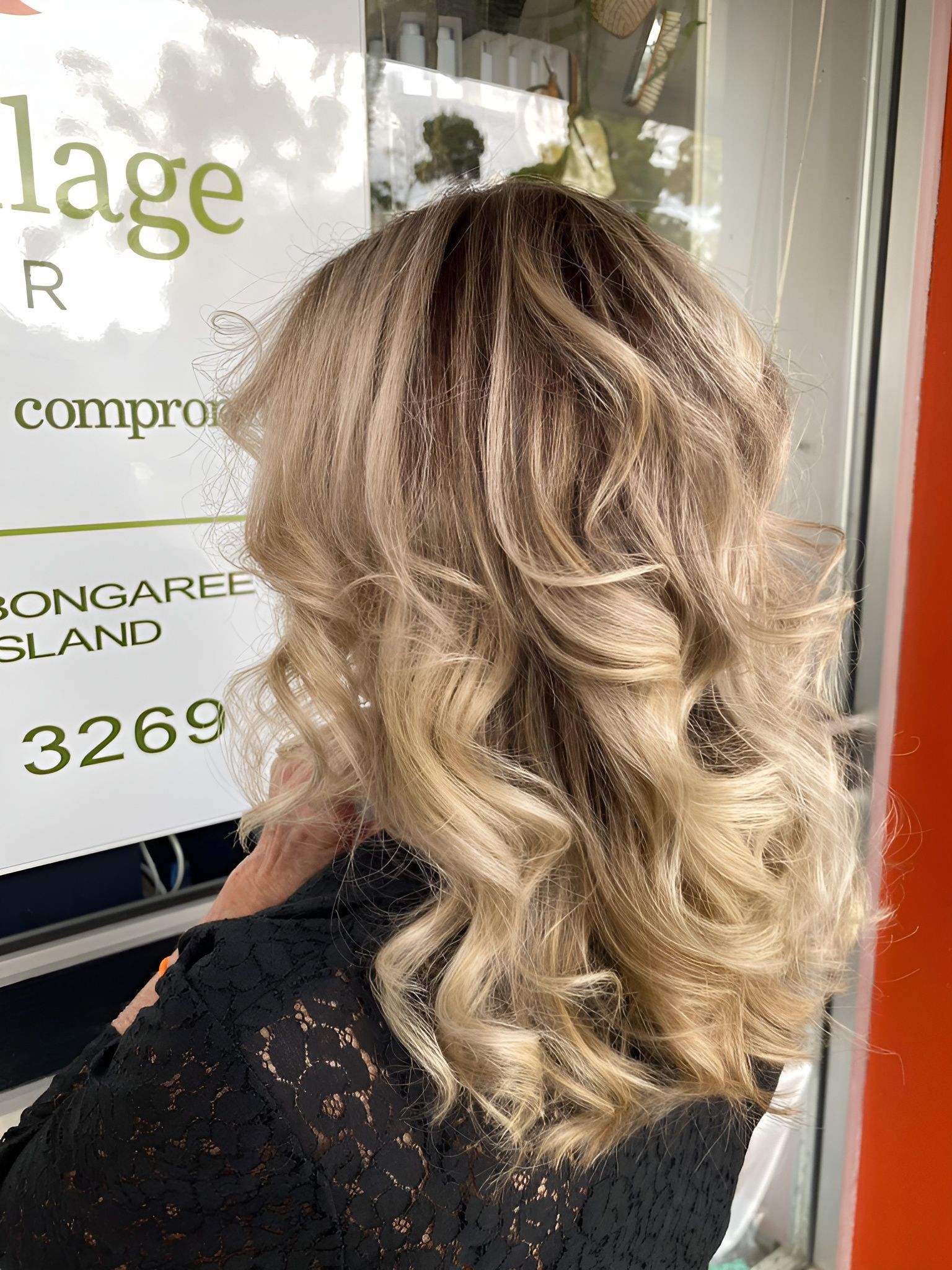 A Woman with Blonde Curly Hair Is Standing in Front of A Sign — Eco Village Hair in Bongaree, QLD
