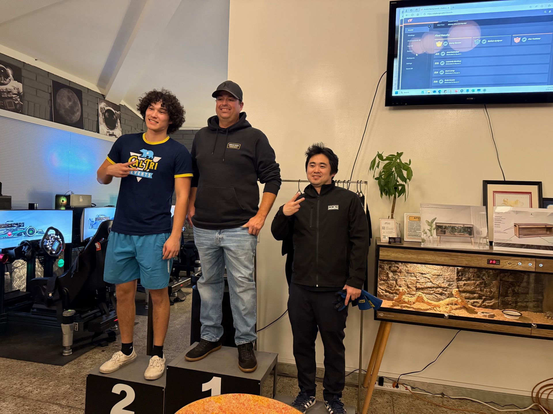 Podium with three people: two men and a young man. The young man is making a hand gesture, the other two are smiling. A display is behind them.