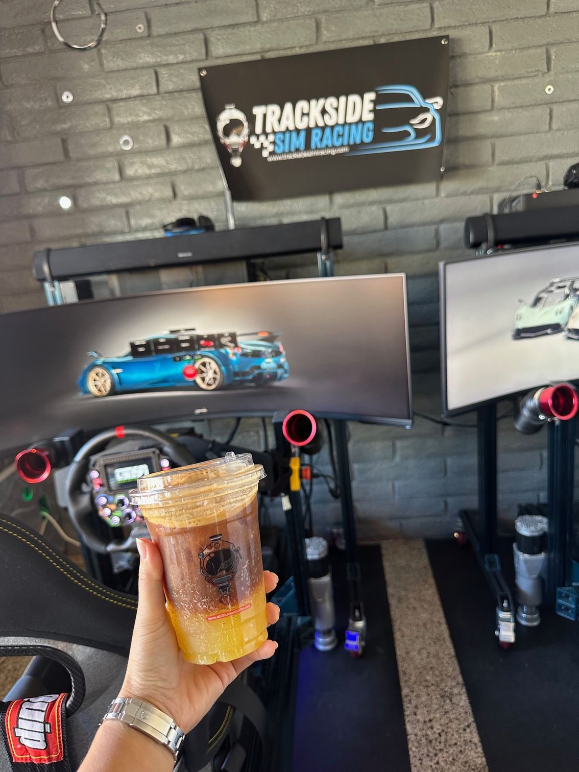 A hand holding an iced coffee in front of a racing simulator setup.