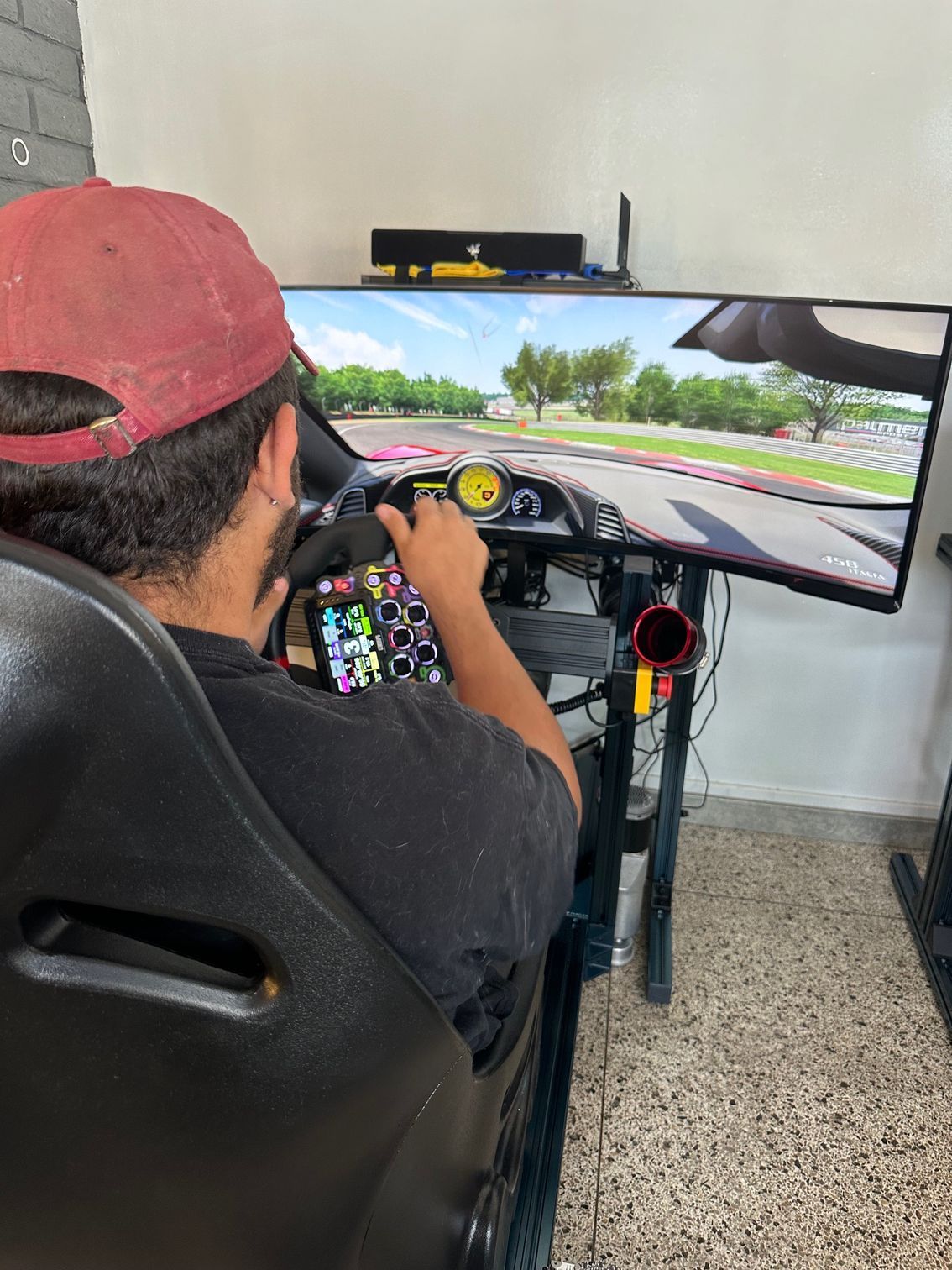 Person in a racing simulator with a red cap, playing a game on a large screen.