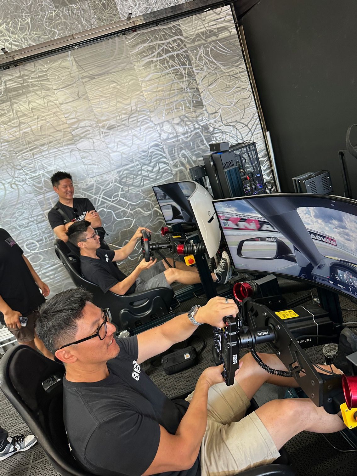 Men at racing simulator stations, one driving, others watching, in a brightly lit room.