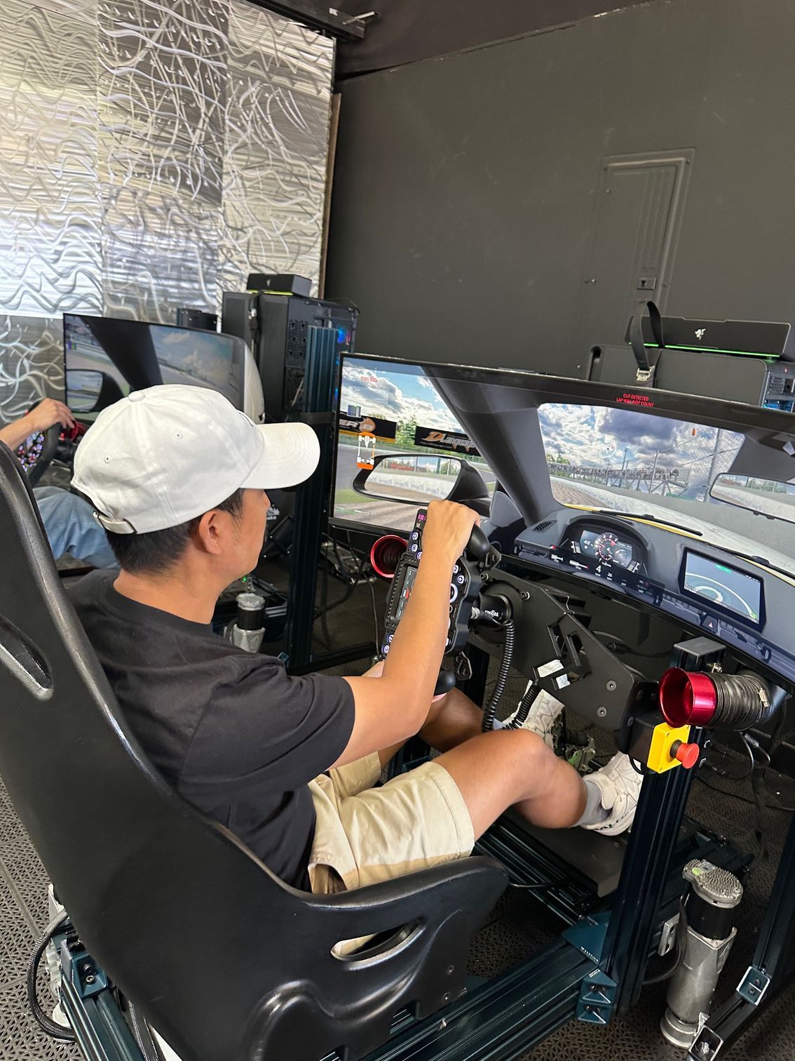 Man in a baseball cap using a racing simulator. He's gripping the steering wheel, looking at screens.