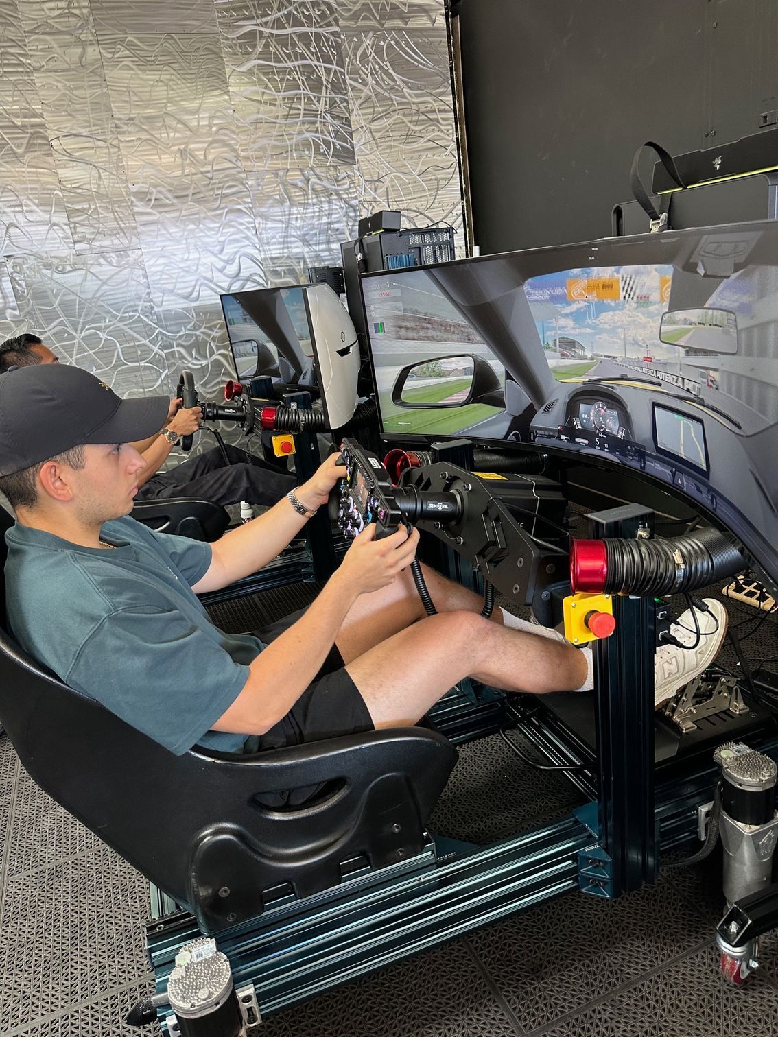 Man in racing simulator, gripping steering wheel, focused on the screen. Other simulator in background.