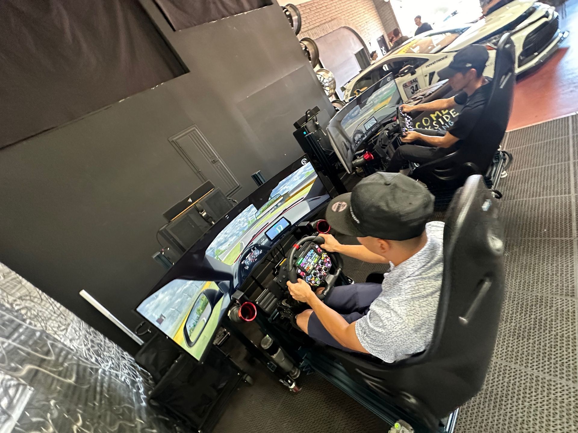 Two men using racing car simulators, both wearing hats and focused on the screens in a workshop.