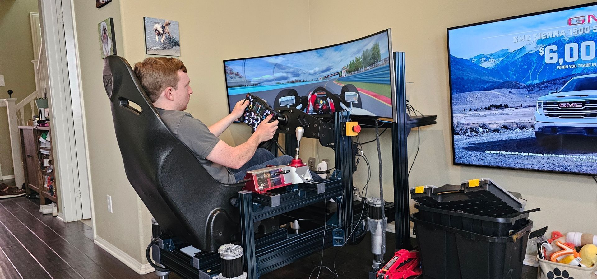 A person sits in a racing simulator, holding a steering wheel. There is a screen showing a racing game.