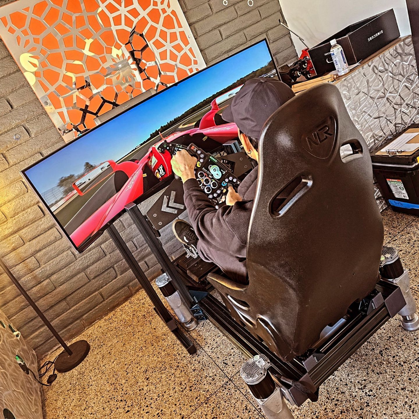 Person using a racing simulator with a large monitor. The room has exposed brick and a colorful art piece.