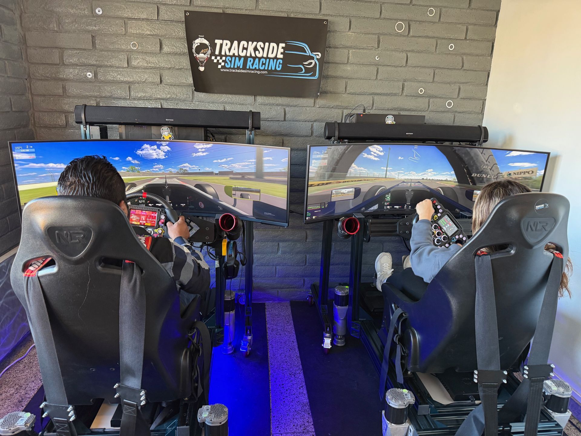 Two people racing on simulated driving game. Inside a room with large screens and racing chairs.