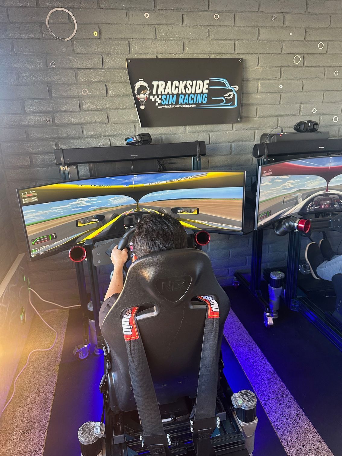 Person racing in a simulator with a steering wheel and monitors at Trackside Sim Racing.