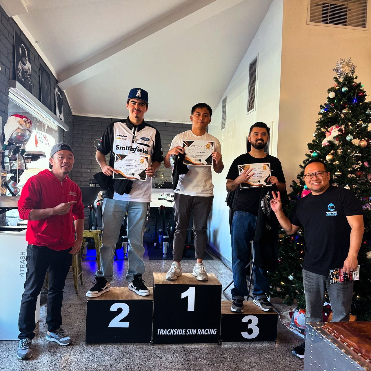 Winners stand on a podium. Three men hold certificates; a Christmas tree is in the background.