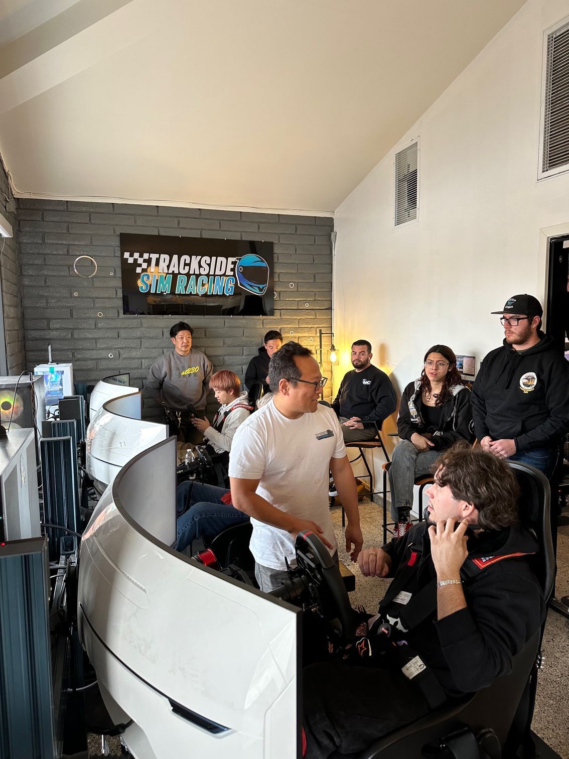 A group of people inside a sim racing stadium. Some seated in racing simulators, others watching. White and black decor.