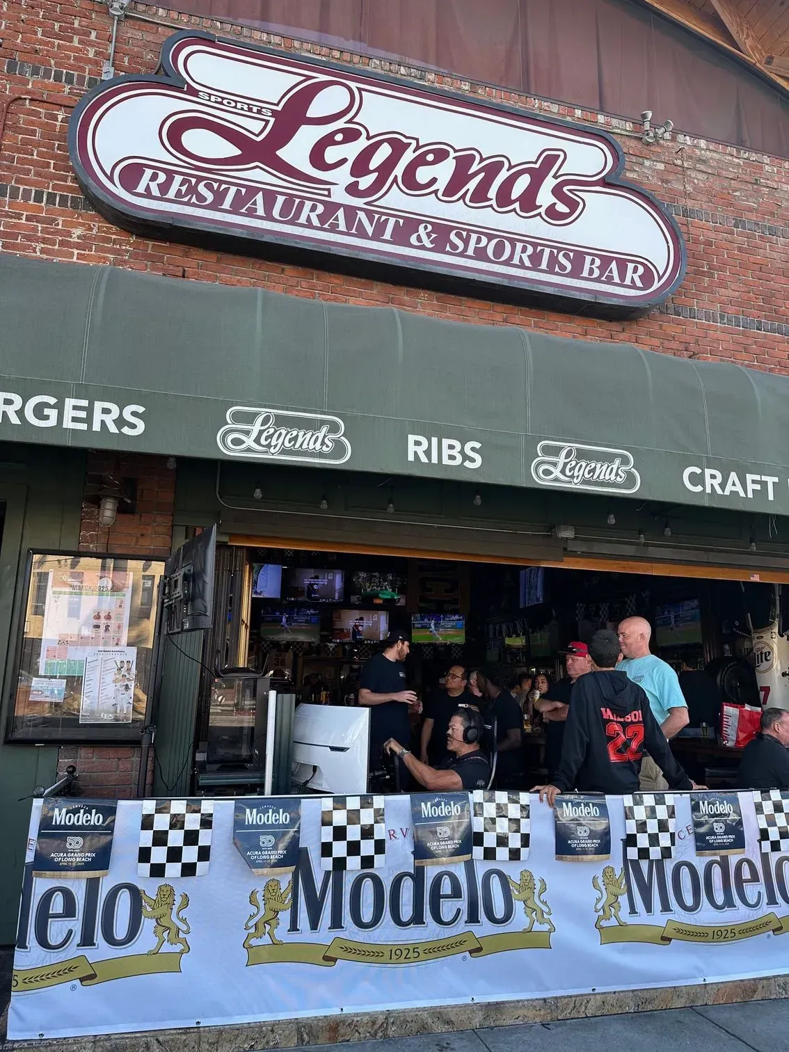 Legends Restaurant & Sports Bar exterior with customers at the bar. A Modelo banner is in front.