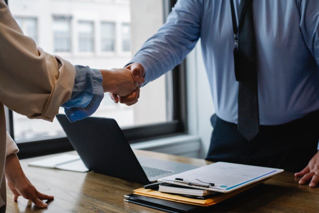 People shaking hands at a desk with a laptop and papers, likely in an office setting.
