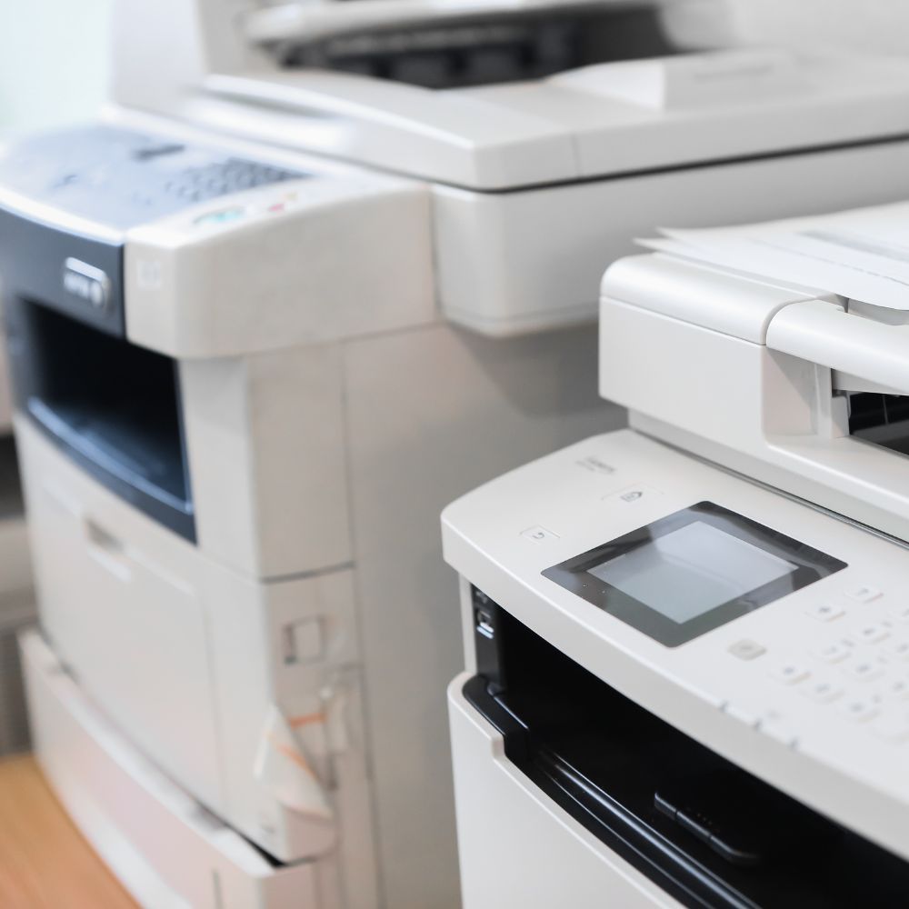 White and gray office printers side-by-side, with control panels and paper trays.