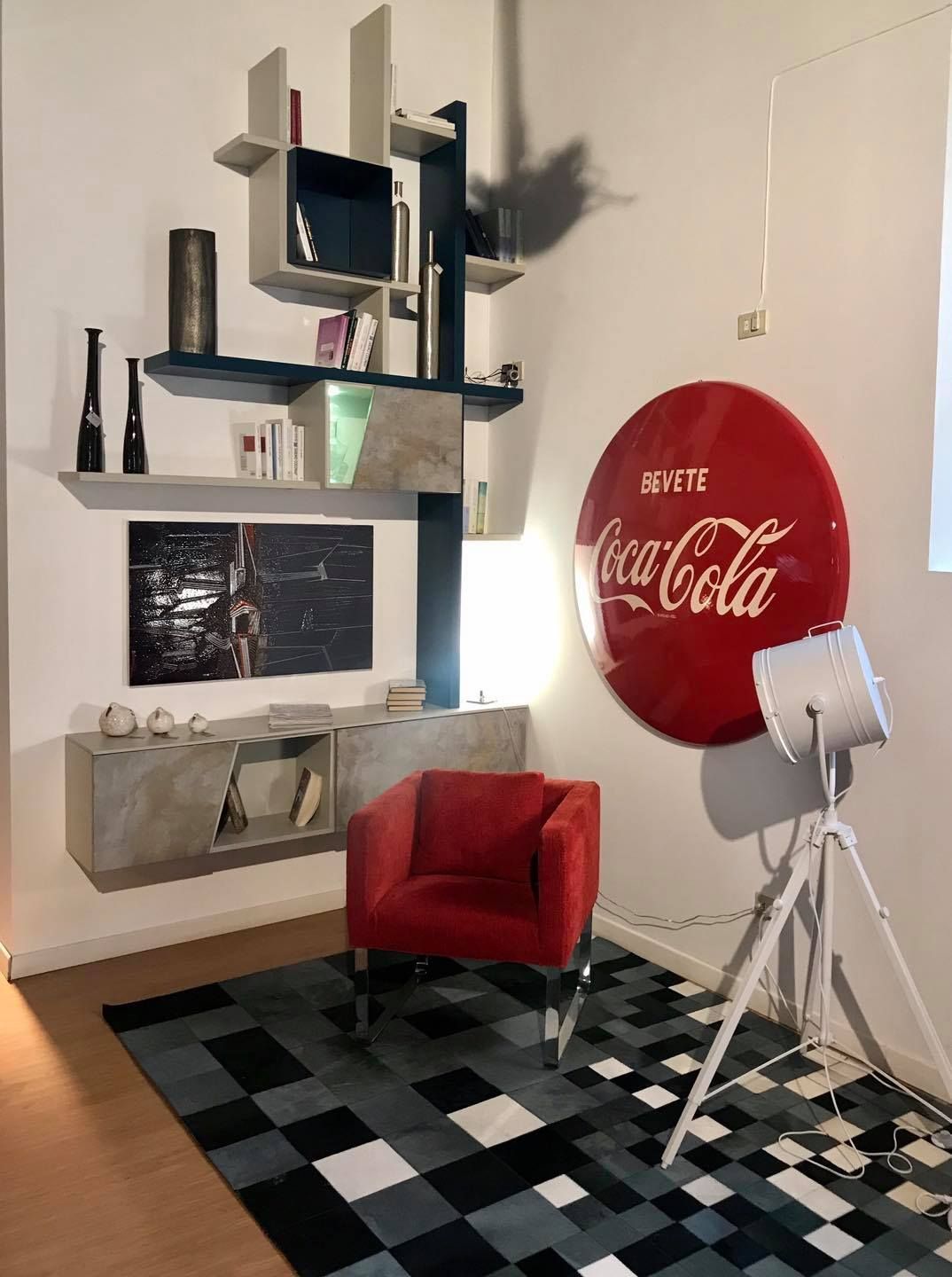 Soggiorno con insegna della Coca-Cola, scaffali moderni, sedia rossa, tappeto geometrico e faretto.