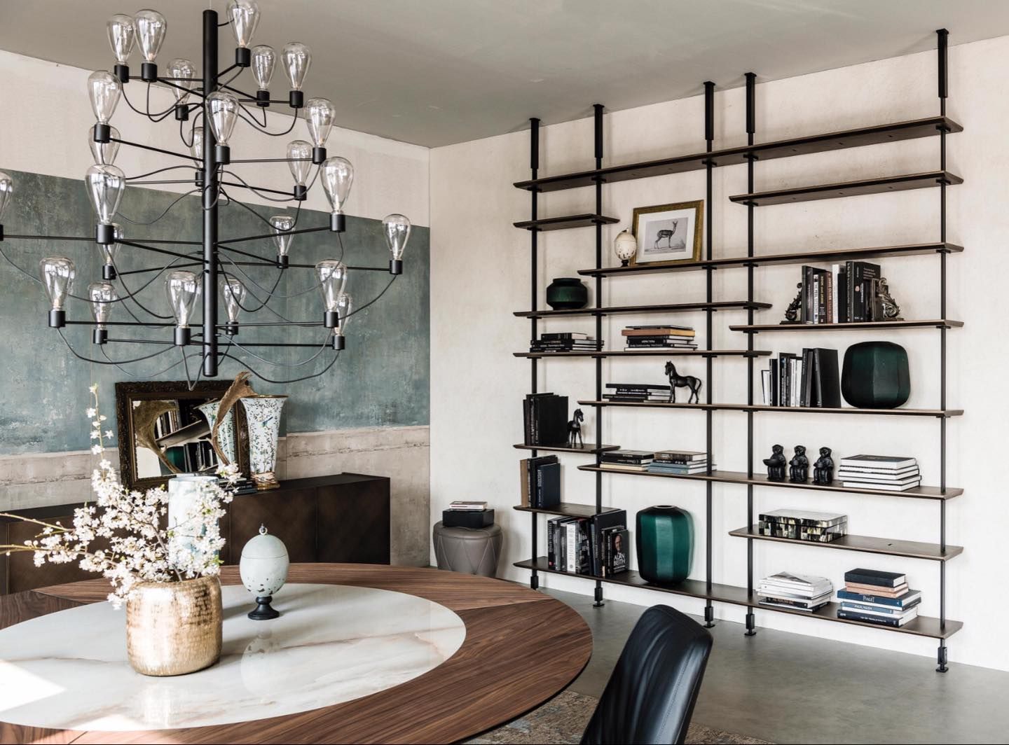 Sala da pranzo con tavolo rotondo, grande lampadario e alta scaffalatura nera su cui sono esposti libri e decorazioni.