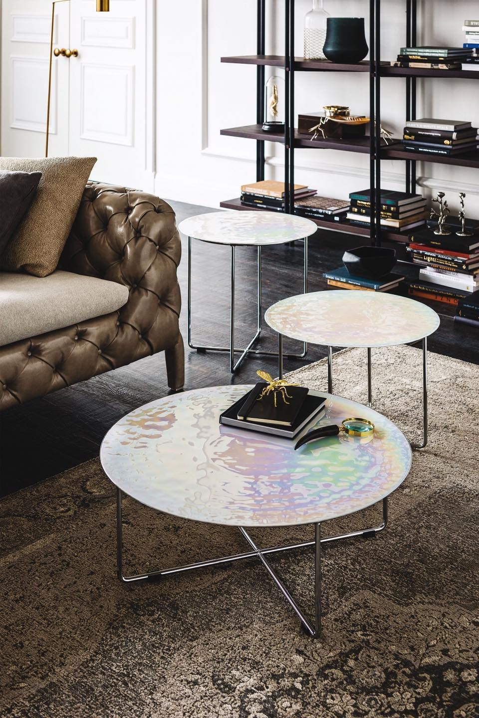 Tre tavolini da caffè con piano in vetro iridescente e gambe cromate su un tappeto fantasia vicino a un divano capitonné e a una libreria.