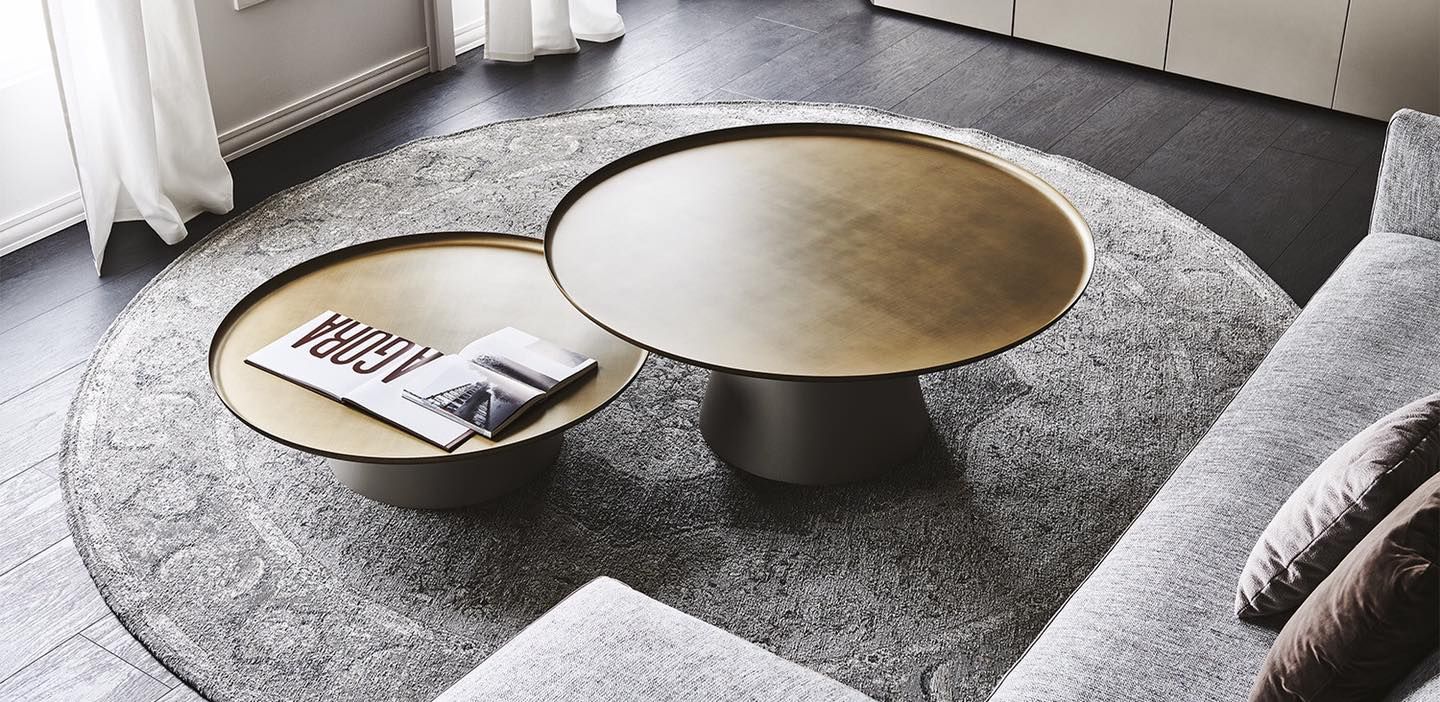 Un tappeto grigio rotondo con due tavolini da caffè con piano dorato e un divano grigio.