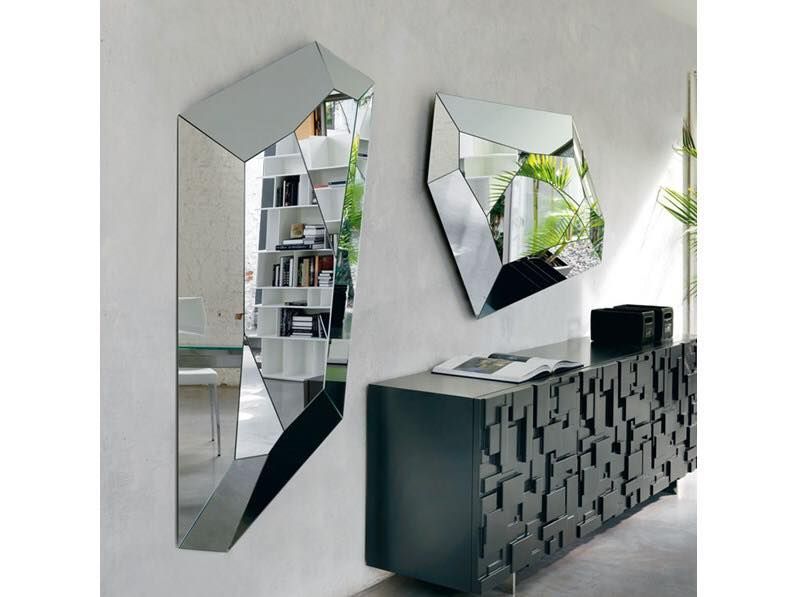 Due specchi geometrici appesi sopra un mobile scuro riflettono una libreria e del verde contro una parete bianca.