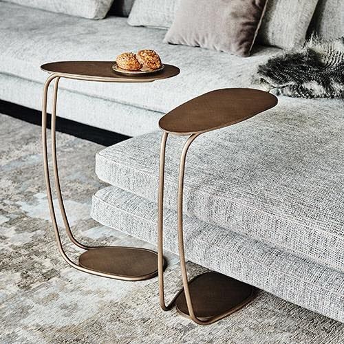 Due tavolini laterali in bronzo a forma di C, ciascuno con un piano ovale, accanto a un divano grigio.