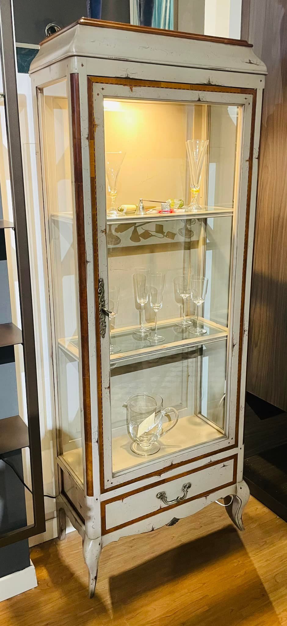 Una vetrina alta e decorata con ripiani in vetro, contenente oggetti decorativi in ​​vetro, illuminata dall'interno.