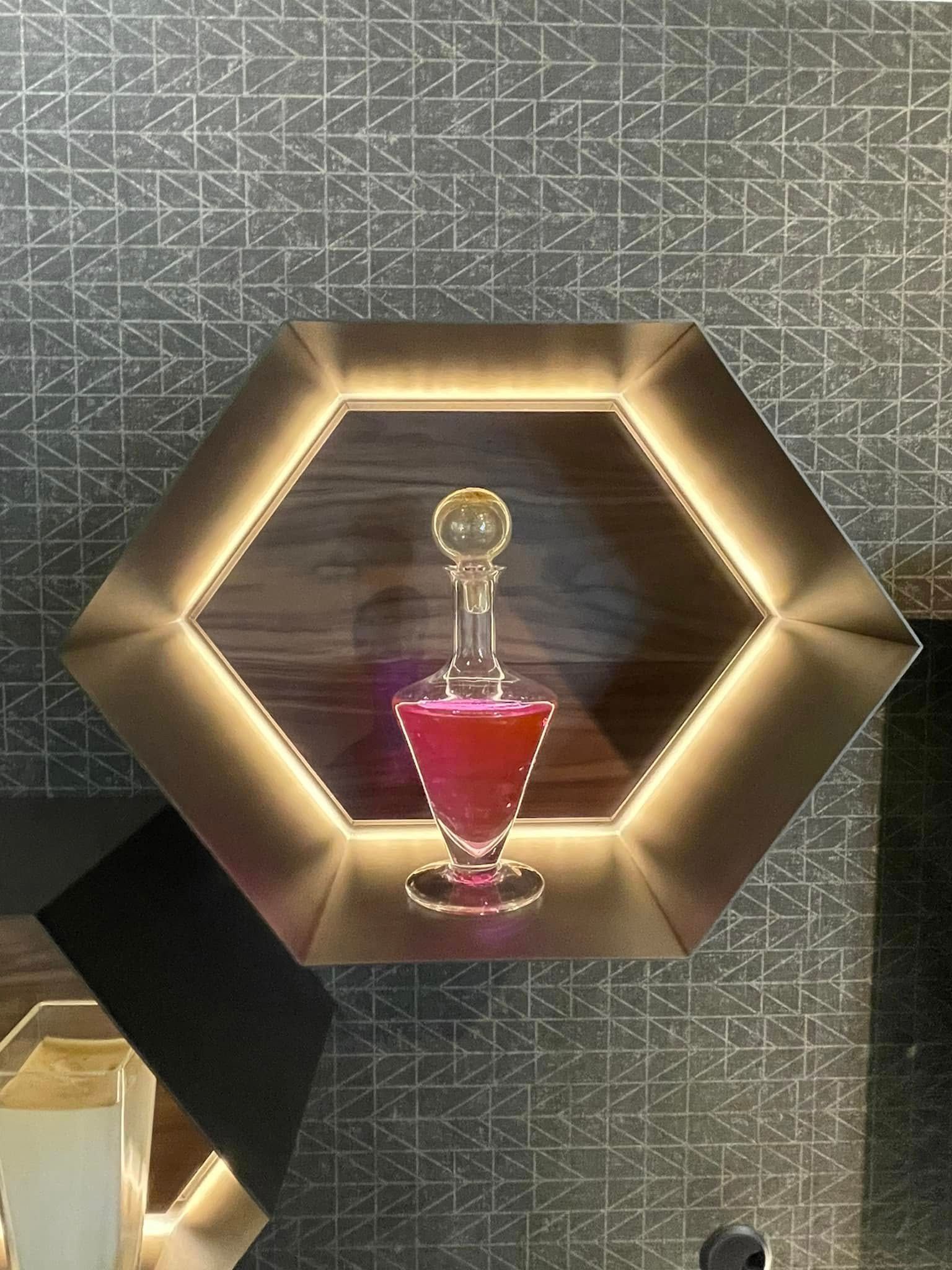 Mensola esagonale con bordi illuminati, su cui è esposta una bottiglia di profumo rossa contro una parete decorata.