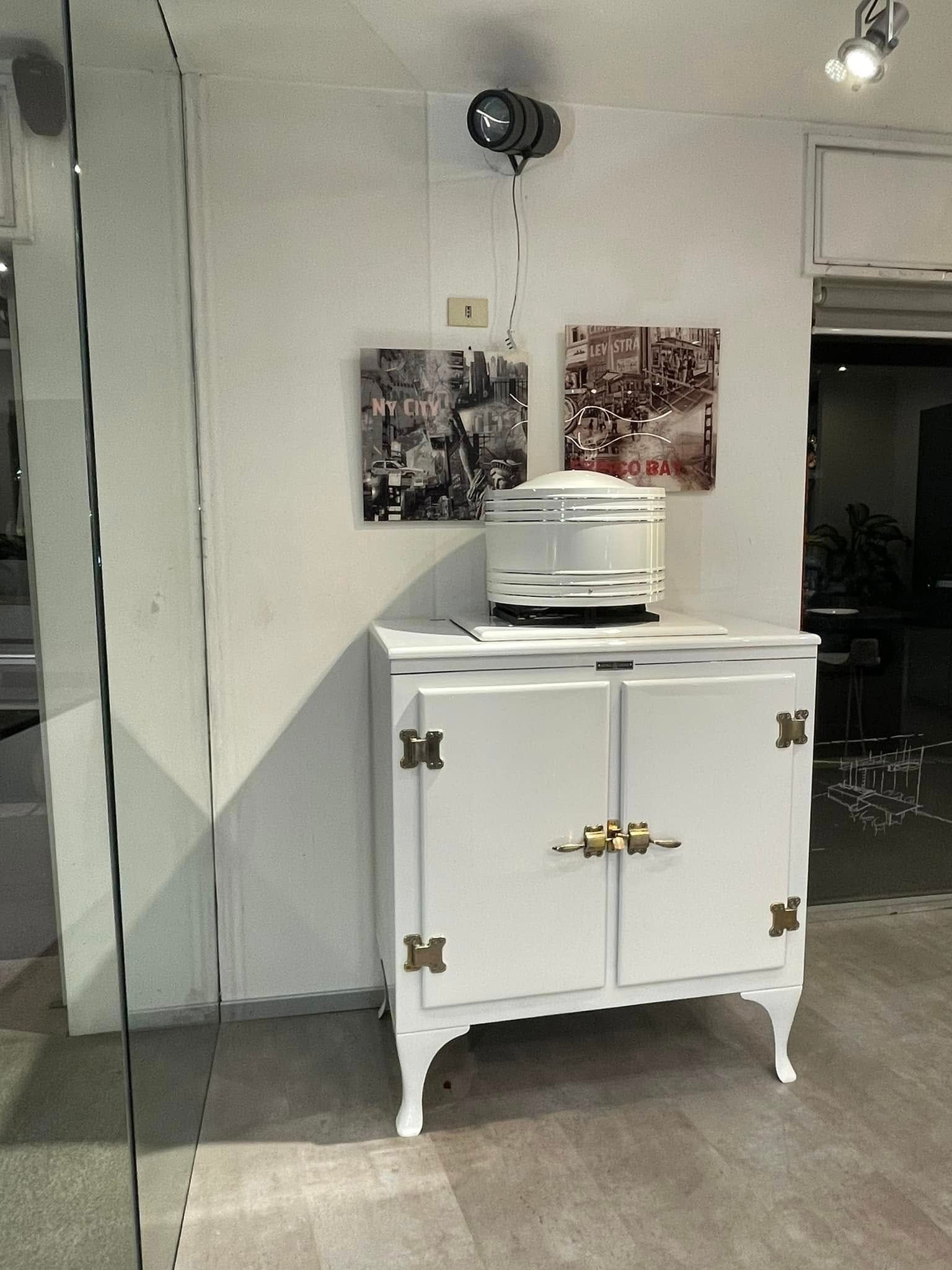 Mobile bianco con ferramenta dorata, contenente un elettrodomestico bianco. Opera d'arte alla parete, faretto sopra.