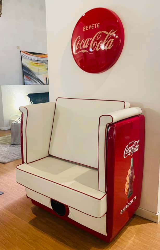 Sedia rossa a tema Coca-Cola con cuscini bianchi, sopra la quale è appesa un'insegna circolare della Coca-Cola.