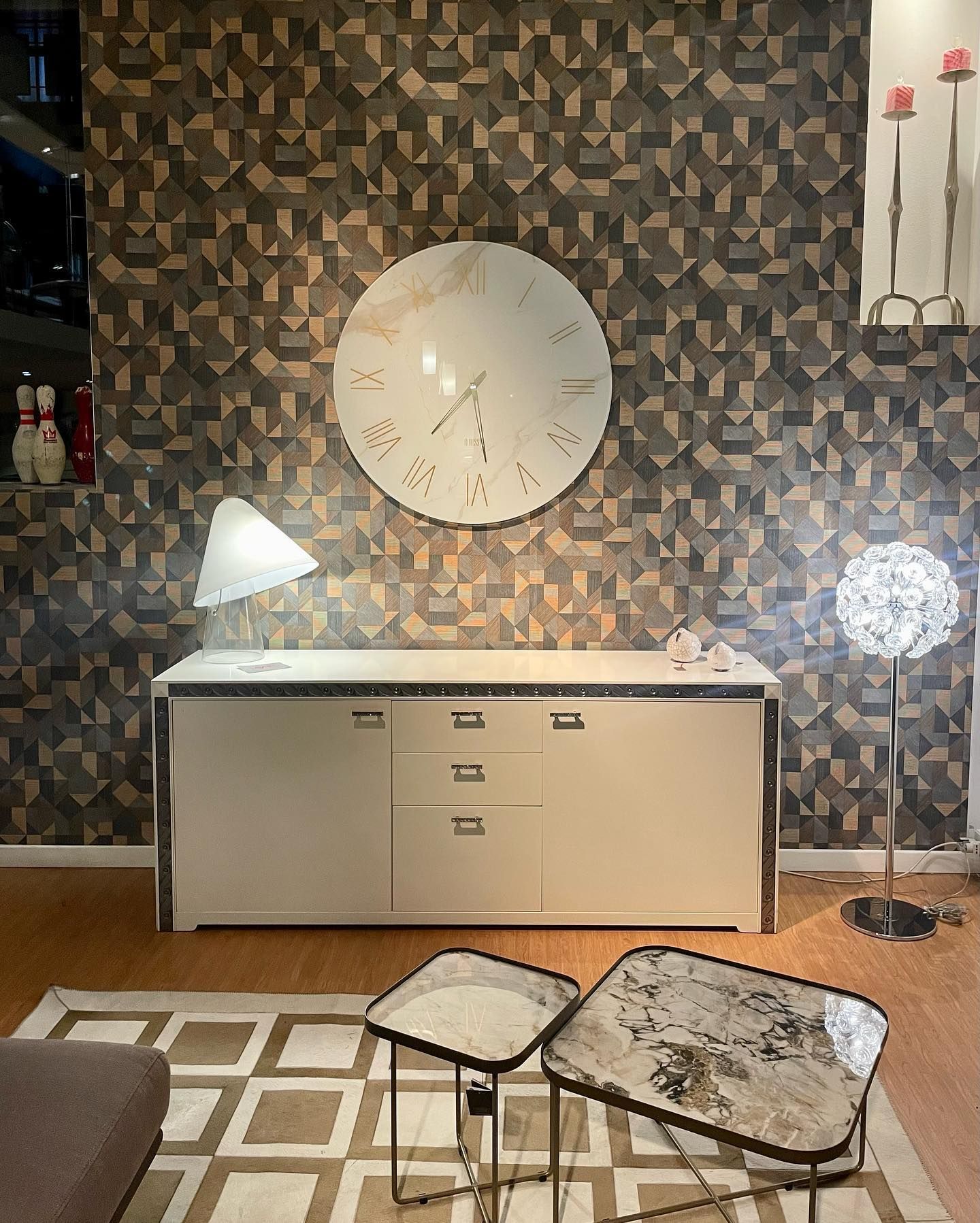 Un soggiorno con una credenza color crema, un orologio, lampade e due tavolini su un tappeto geometrico.