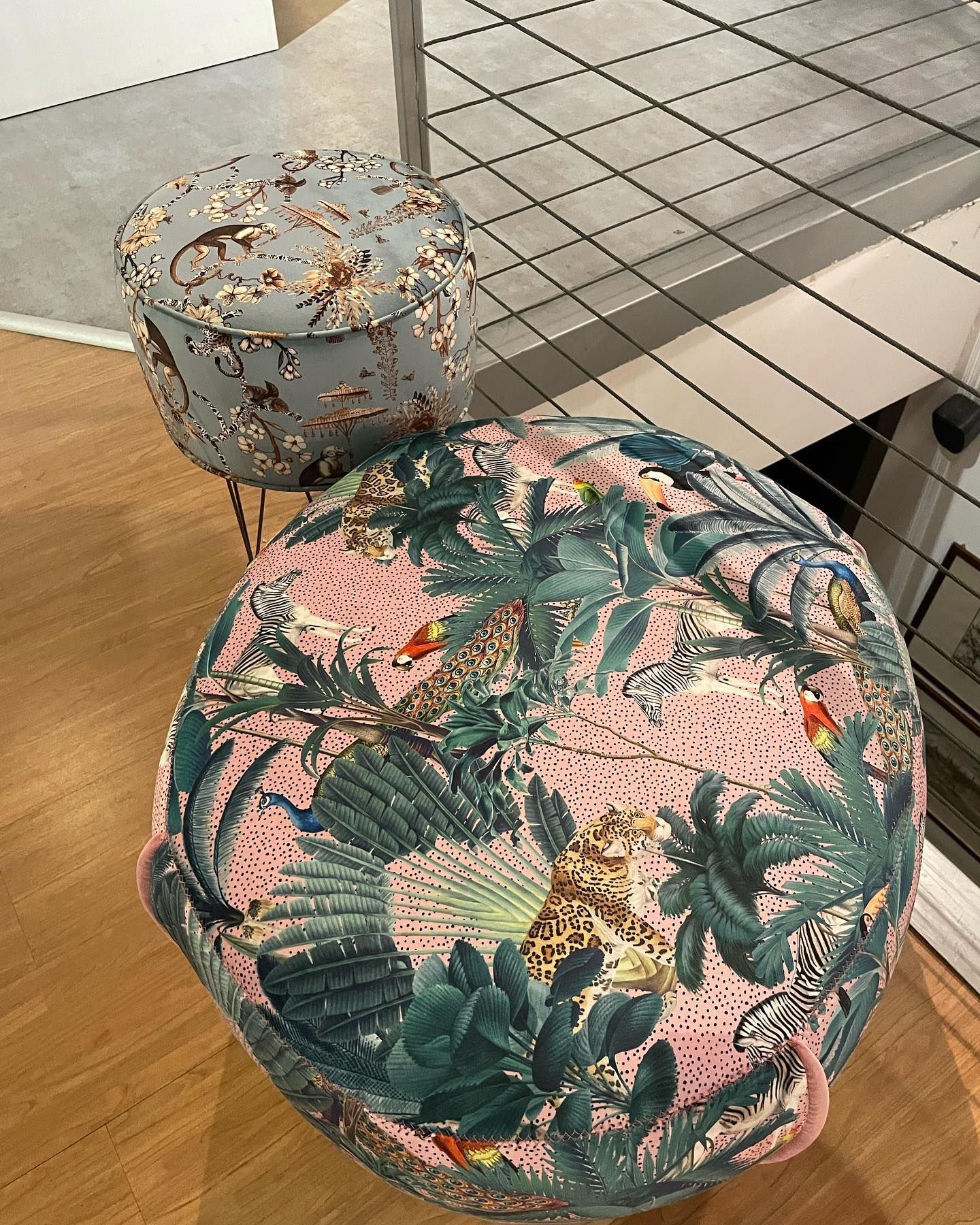 Due pouf decorati, uno grigio con animali, l'altro rosa con foglie della giungla, accanto a una ringhiera di metallo.