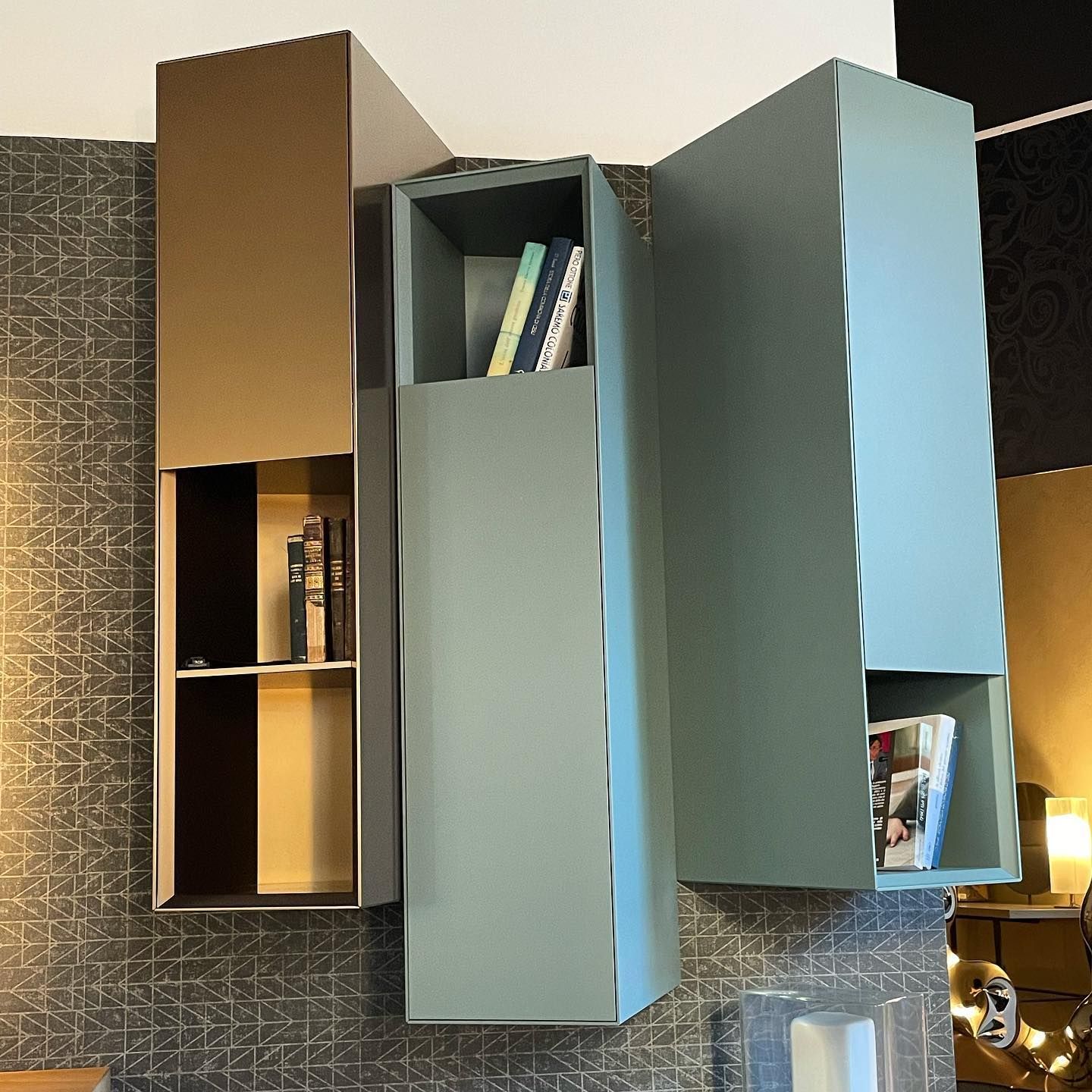 Tre mensole colorate, asimmetriche e montate su una parete decorata, con libri e oggetti.