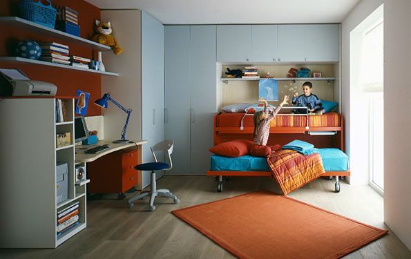 Cameretta per bambini dai colori vivaci con due bambini. Pareti, scrivania, letto e scaffali arancioni e blu. Un bambino in piedi, l'altro seduto.